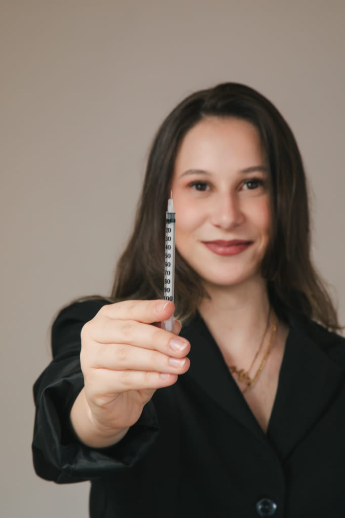 Dra. Júlia de blazer preto segurando uma seringa em primeiro plano