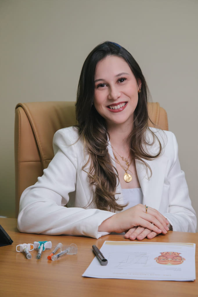 Dra. Júlia Laguna Dumke sorrindo, de blazer branco, sentada à mesa